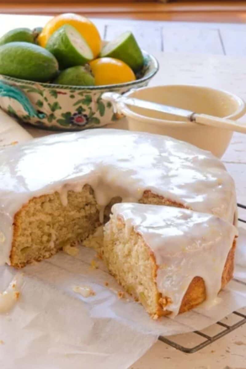 A moist, freshly baked cake with a creamy white glaze on top, with a bowl of whole feijoas in the background, suggesting a key ingredient.