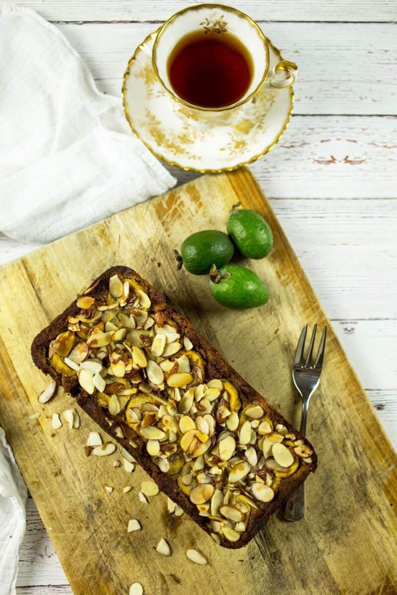 An overhead view of a feijoa and banana loaf on a wooden board, sliced to reveal the moist interior, topped with slivered almonds, next to a vintage cup of tea.