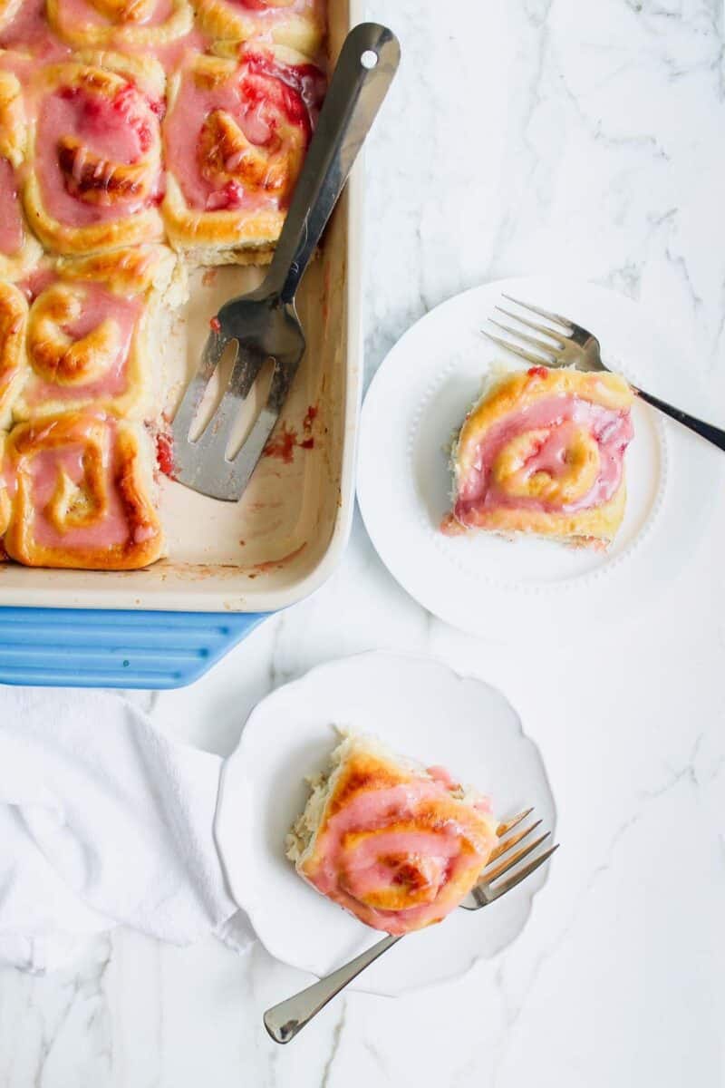 Pink rhubarb rolls in a blue baking dish, with a serving plate showing a frosted roll beside it.