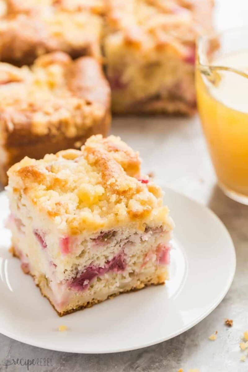 A slice of a layered coffee cake with a crumb topping on a white plate, next to a glass of orange juice. The cake has chunks of rhubarb within, offering a sweet and tart flavor combination.