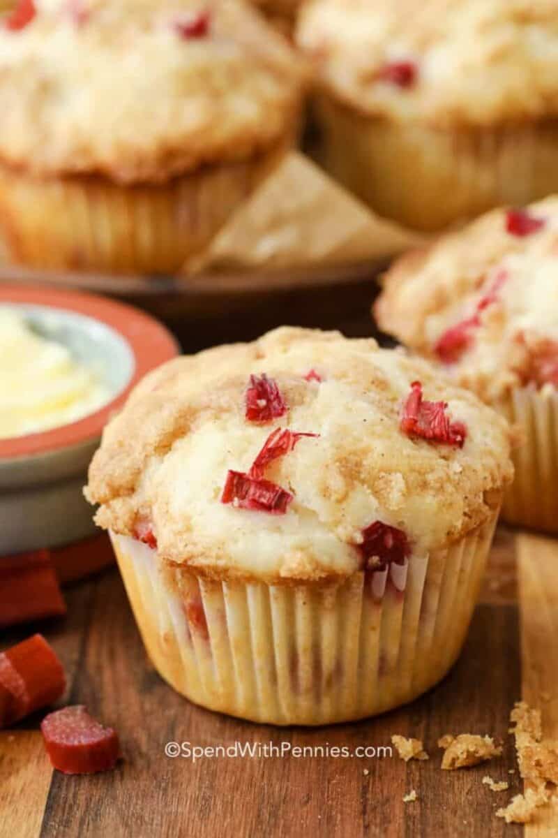 Rhubarb muffins with a golden-brown crumb topping, showcased on a wooden surface. The muffins have visible pieces of red rhubarb peeking out, and there's a pat of butter in the background.