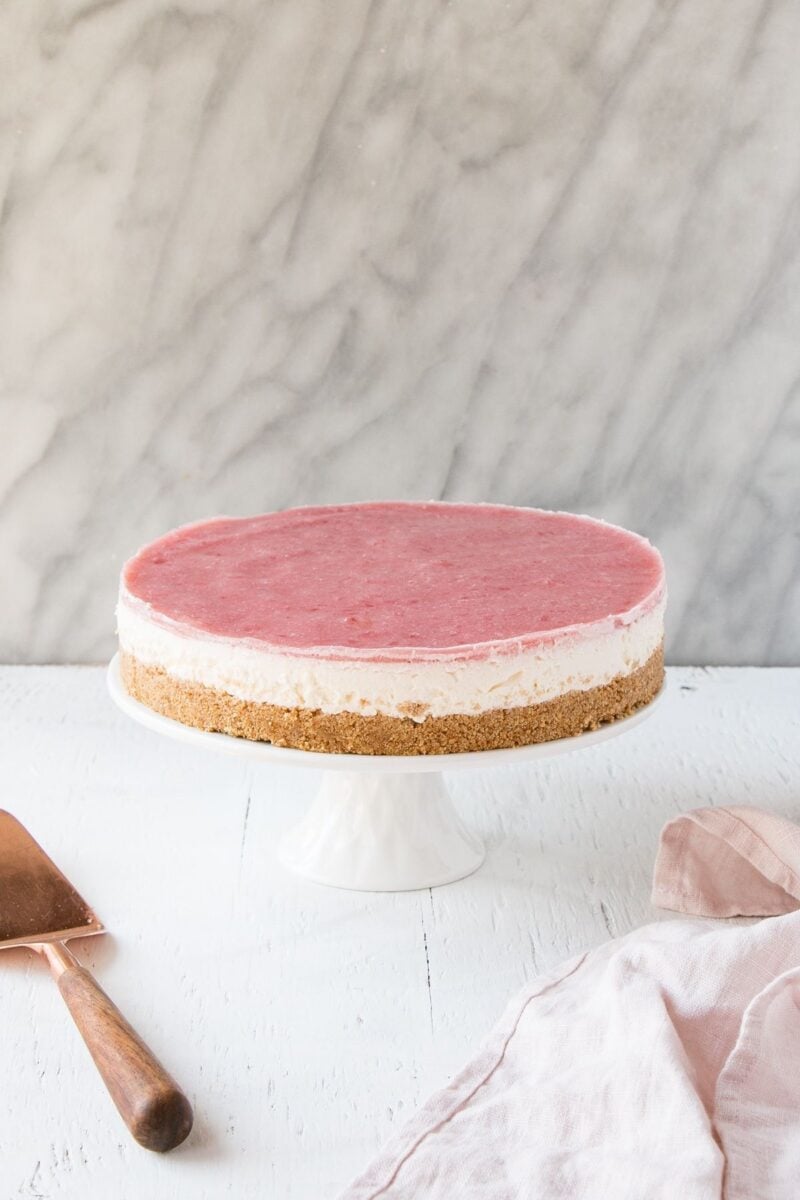 A no-bake cheesecake with a thick layer of rhubarb gel on top, set on a classic cream cheese filling. The cheesecake has a crumbly base and is placed on a white cake stand, with a pale pink fabric and a cake server alongside.