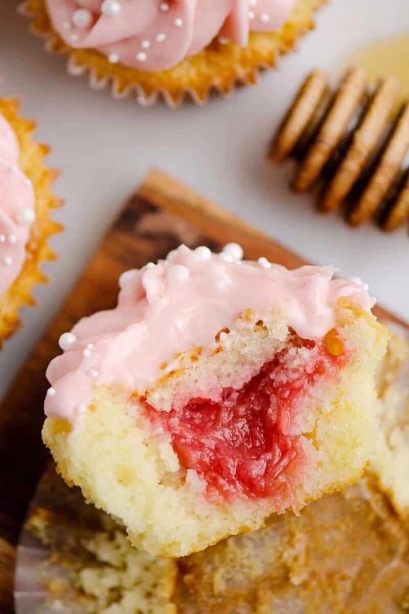 A close-up of a cupcake with pink frosting and white decorative pearls on top. The cupcake is cut in half, revealing a tender crumb and a generous filling of pink rhubarb.