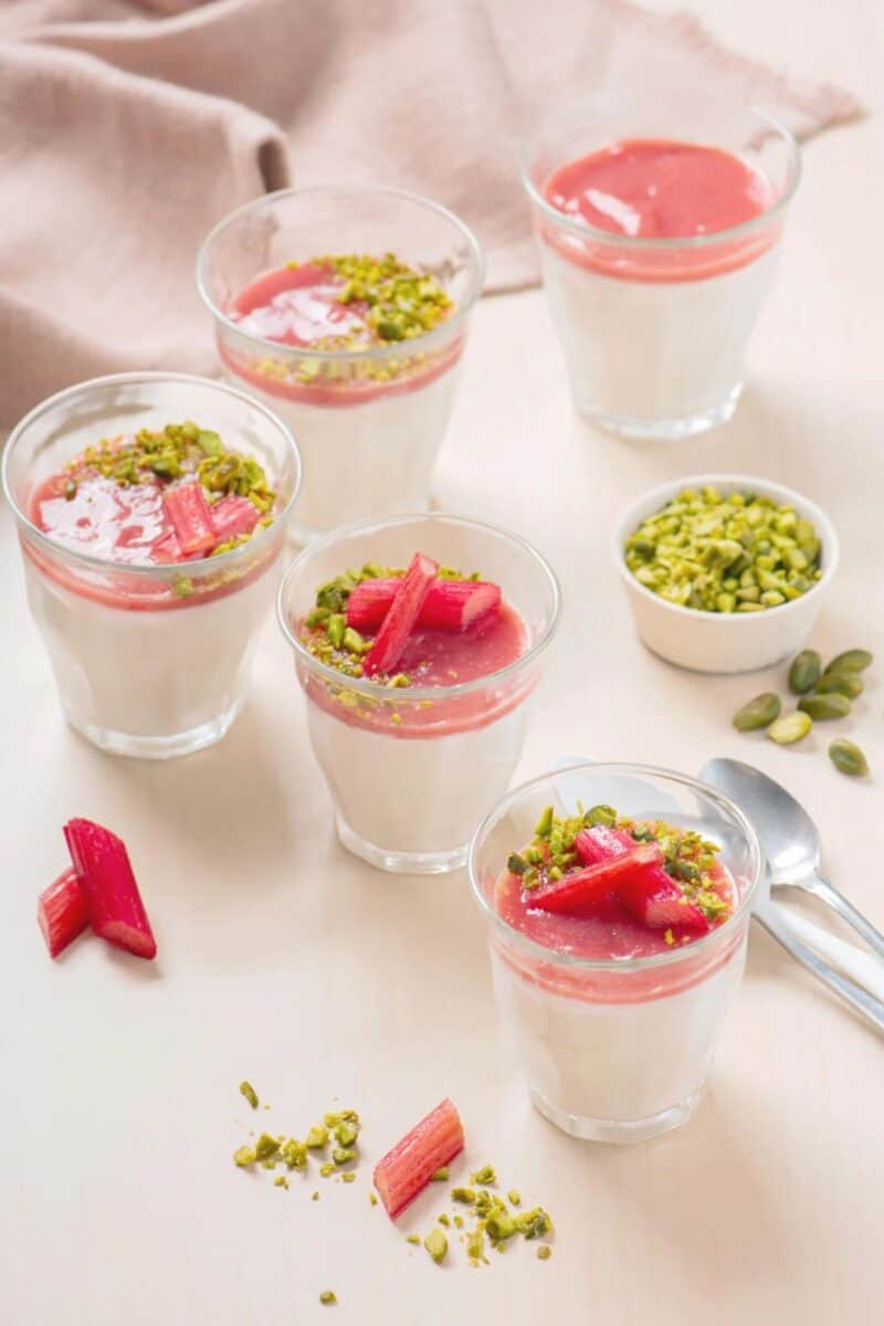 A creamy panna cotta dessert in clear glasses, topped with a layer of pink rhubarb compote and sprinkled with chopped green pistachios, creating a contrast of colors and textures.