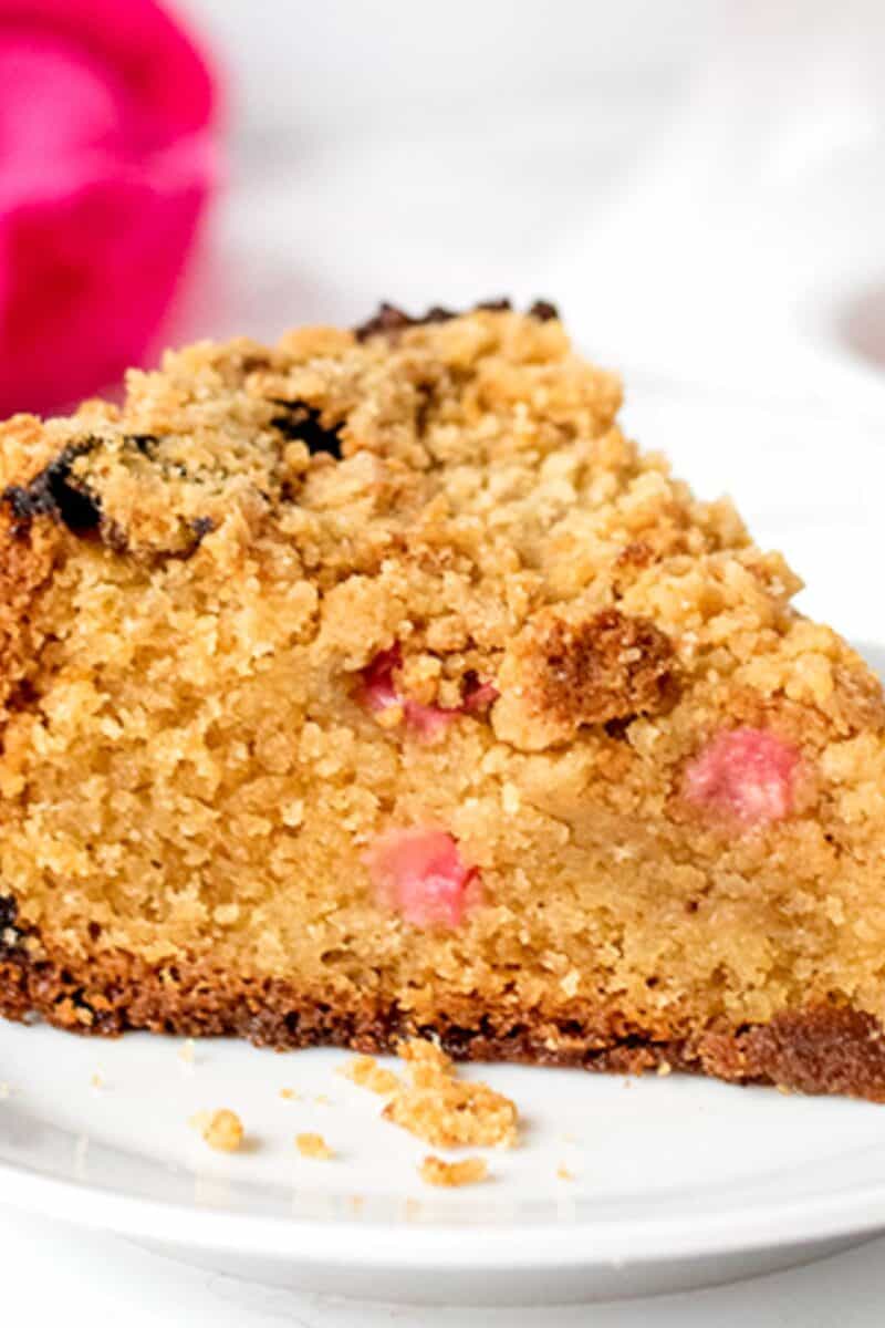 A moist, golden brown coffee cake with a crumbly topping. Visible within the cake are vibrant pieces of pink rhubarb, adding a pop of color and indicating a tart flavor profile.