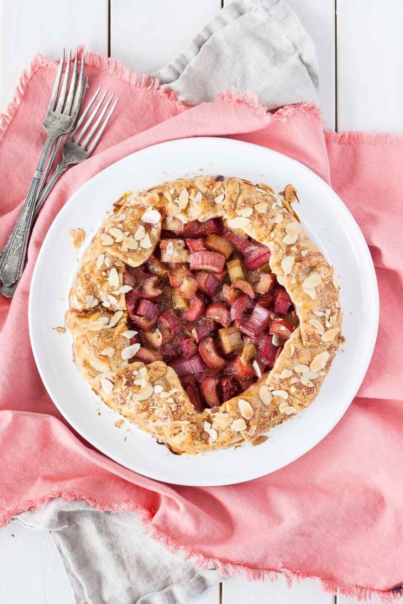 Rustic rhubarb galette with a golden crust sprinkled with sliced almonds, centered on a white plate with a coral napkin.