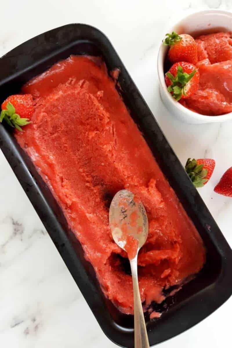 Homemade strawberry rhubarb sorbet in a black loaf pan with a silver spoon and fresh strawberries on the side