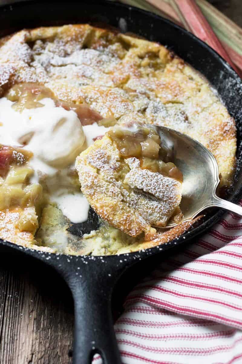 Freshly baked rhubarb Dutch baby pancake dusted with powdered sugar, topped with a dollop of cream, in a cast-iron skillet with a vintage silver spoon
