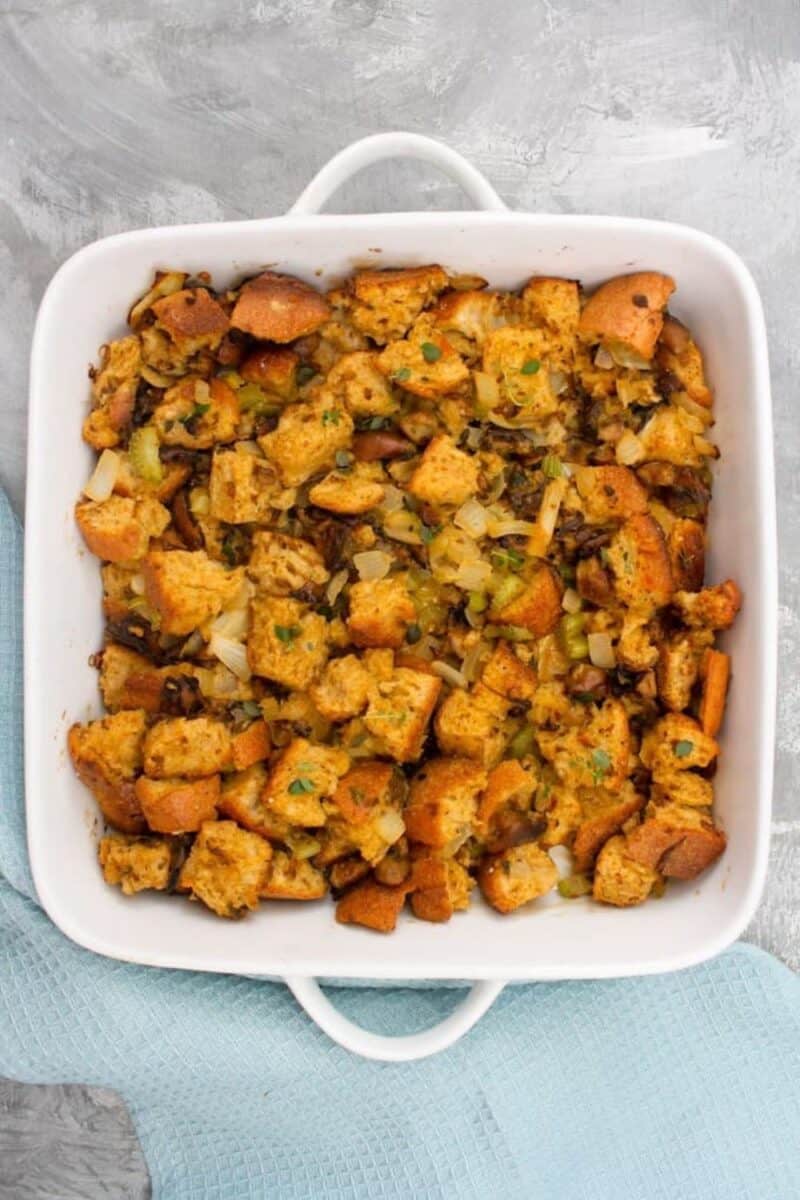 A white casserole dish filled with savory stuffing with chunks of crusty bread, celery, and herbs, presented on a pale blue cloth.