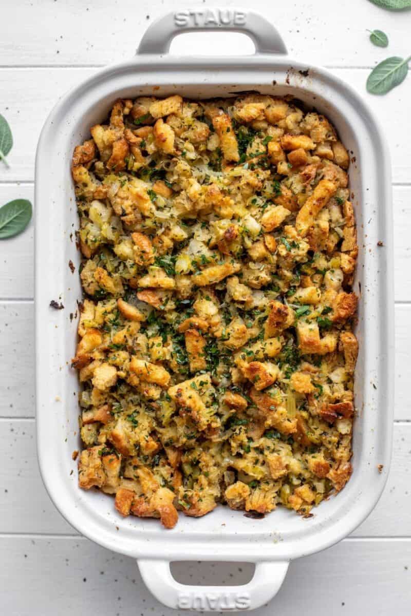 Baked Thanksgiving stuffing in a white ceramic dish, showing crispy golden pieces and fresh herbs on top, on a white wooden background.