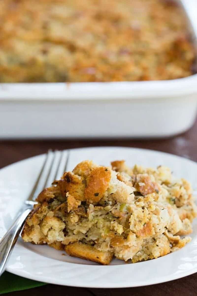 A portion of fluffy Thanksgiving stuffing on a white plate, with visible herbs and a crisp top, beside a full casserole dish.