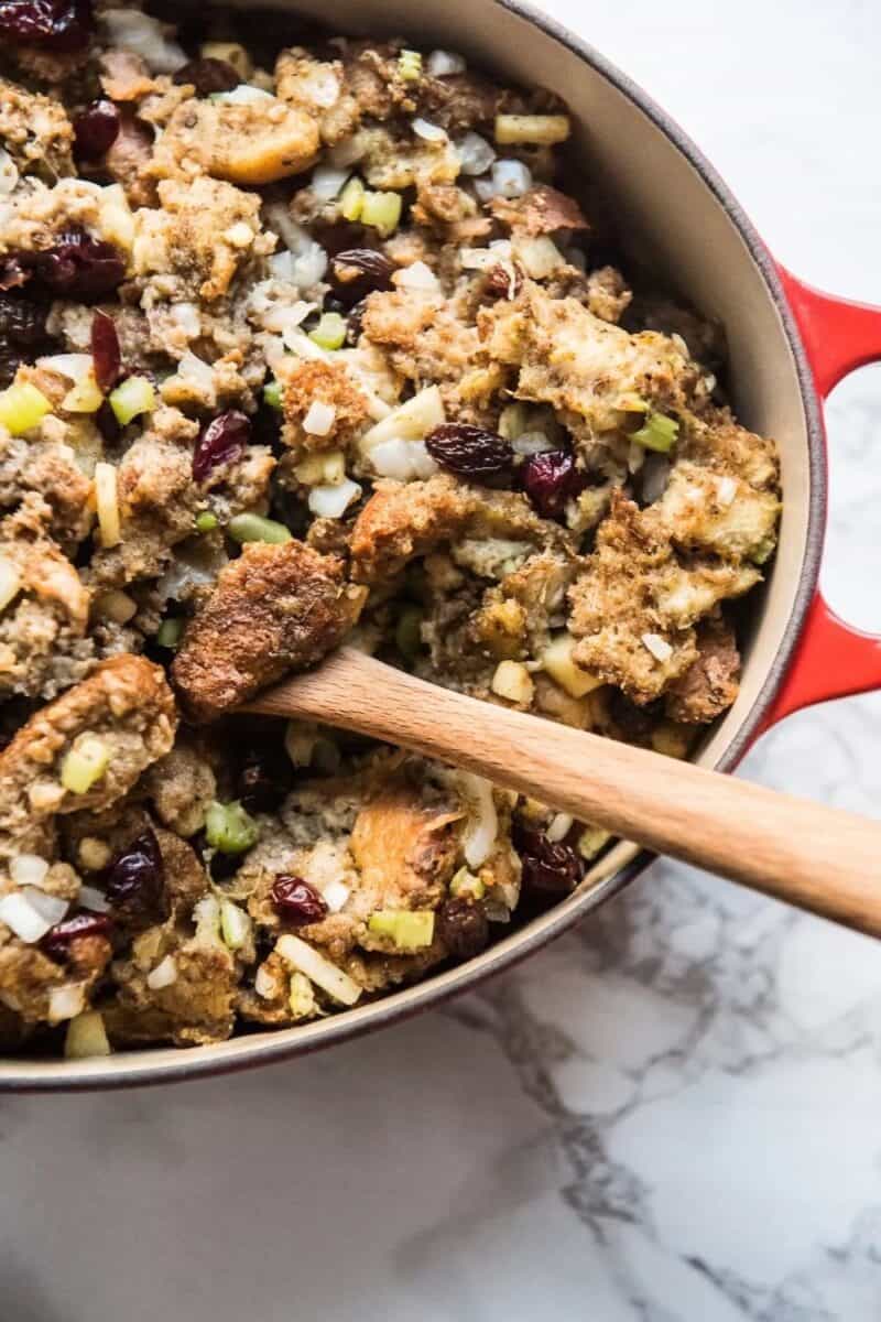 A Dutch oven filled with savory stuffing, featuring dark cranberries, diced apples, and celery, with a wooden spoon.