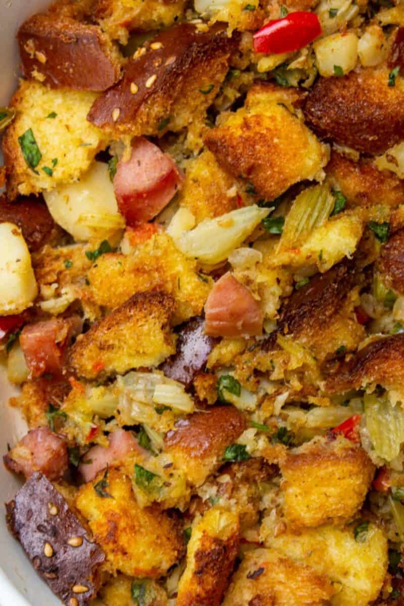 A close-up of vibrant Thanksgiving stuffing in a bowl, with golden cornbread pieces, sausage, and specks of green herbs.
