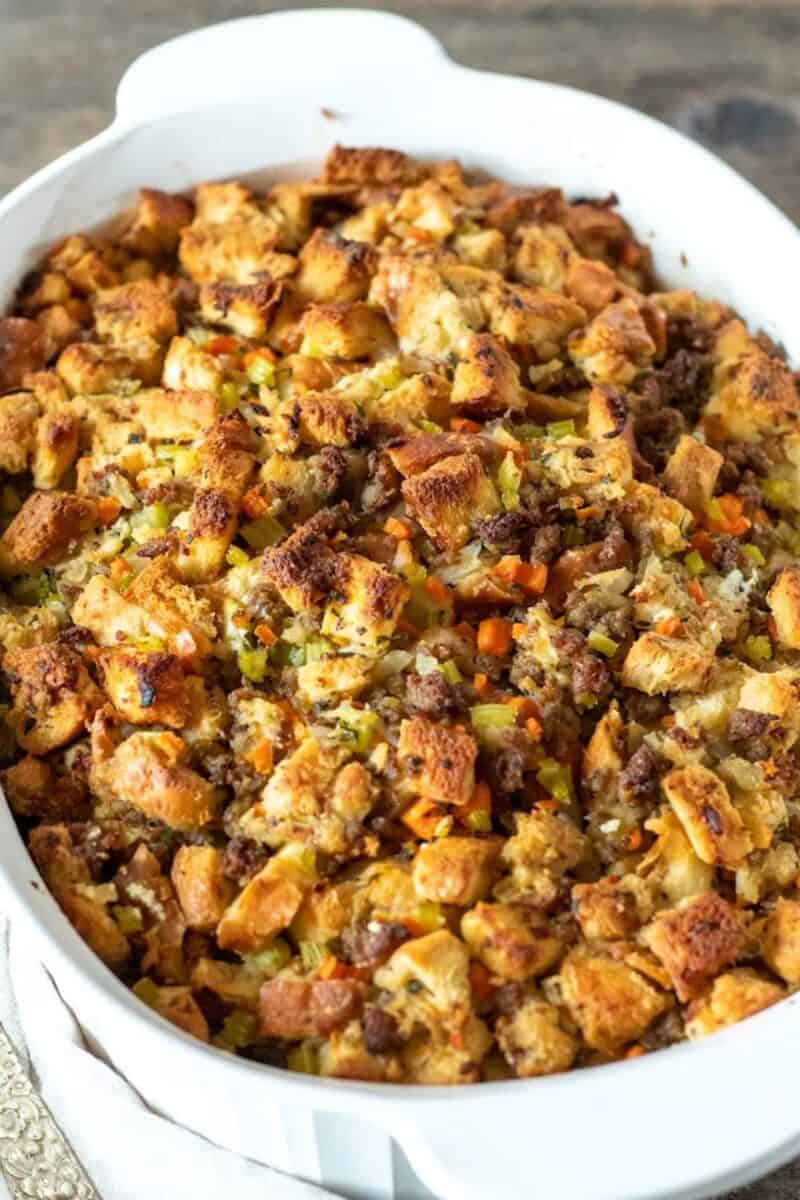 A white casserole dish overflowing with golden-browned stuffing, filled with bits of carrot and celery, with a rustic backdrop.