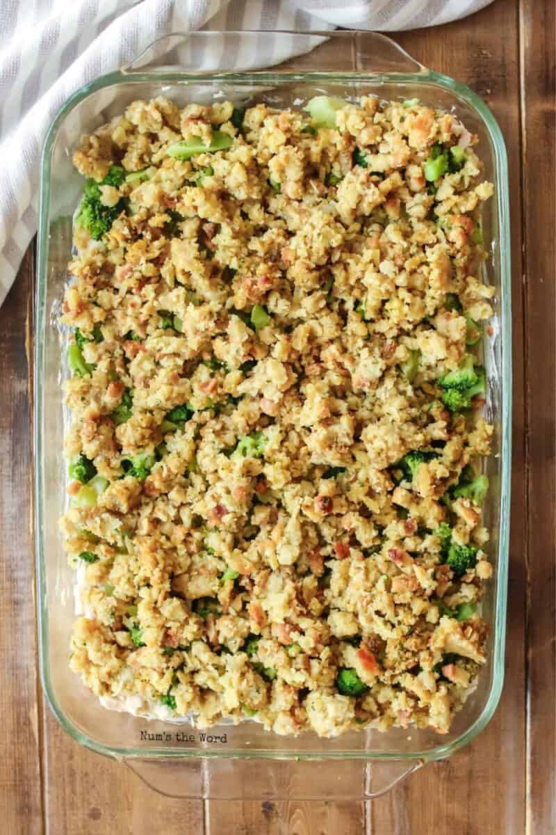 A clear glass baking dish filled with savory bread stuffing mixed with green broccoli and carrots, atop a rustic wooden table.