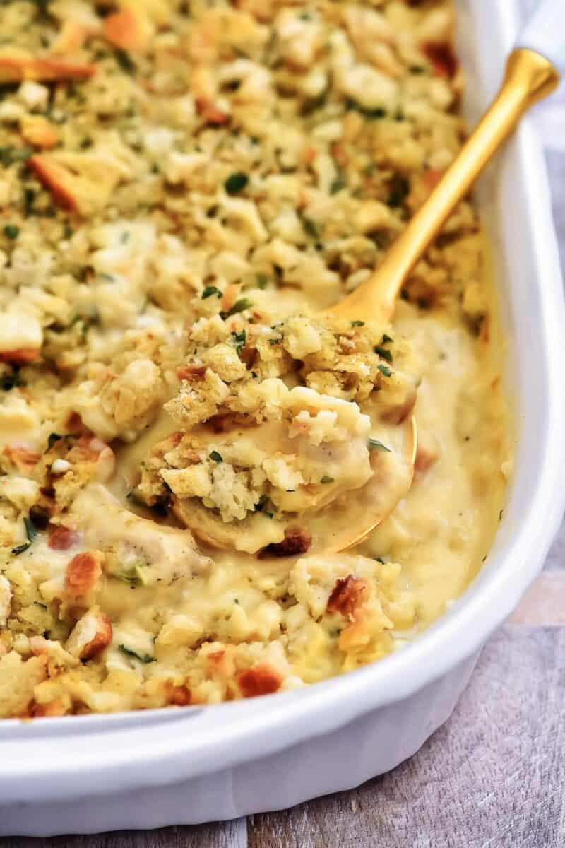 Close-up of creamy Thanksgiving stuffing in a white dish with a golden serving spoon, featuring soft bread chunks and fresh herbs.