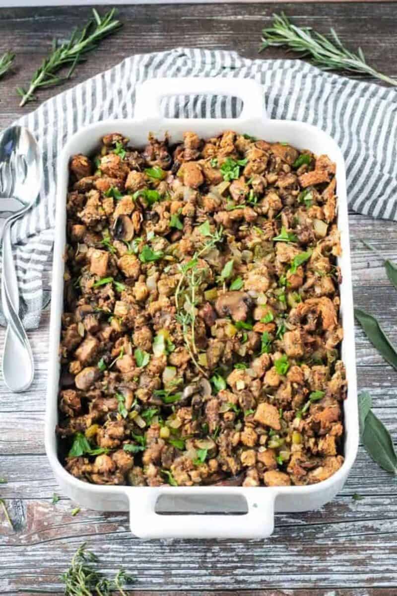 A white casserole dish filled with savory stuffing, garnished with fresh herbs, resting on a striped towel with rustic wooden background.