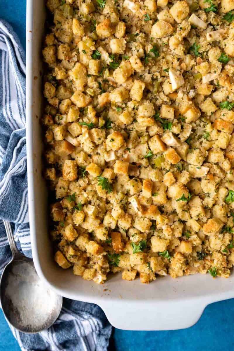 Overhead shot of traditional bread stuffing in a white baking dish, garnished with parsley, alongside a silver spoon on a blue napkin.