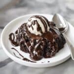A serving of crockpot chocolate lava cake on a white plate, topped with a scoop of vanilla ice cream and drizzled with rich chocolate sauce.