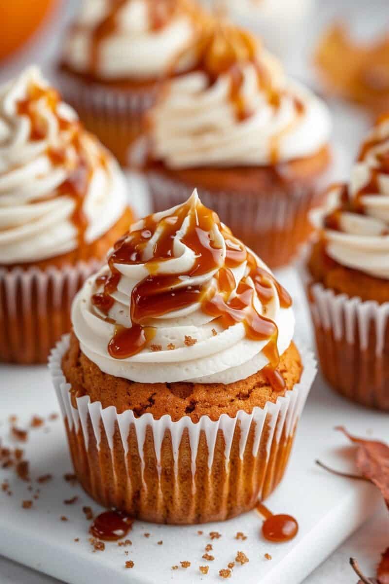 Close-up of Pumpkin Cupcakes with Caramel Cream Cheese Frosting, each topped with a generous drizzle of caramel sauce. The cupcakes are arranged on a white surface, highlighting their moist texture and rich, creamy topping.