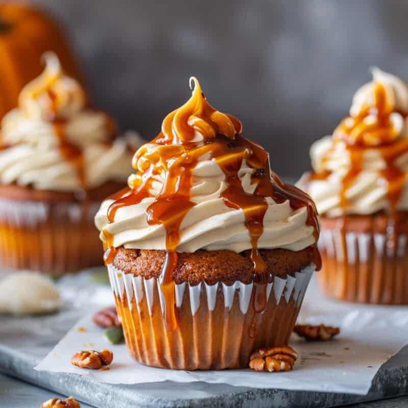 Close-up of Pumpkin Cupcakes with Caramel Cream Cheese Frosting, topped with a generous drizzle of caramel sauce. The cupcakes are arranged on parchment paper, highlighting their moist texture and rich topping.