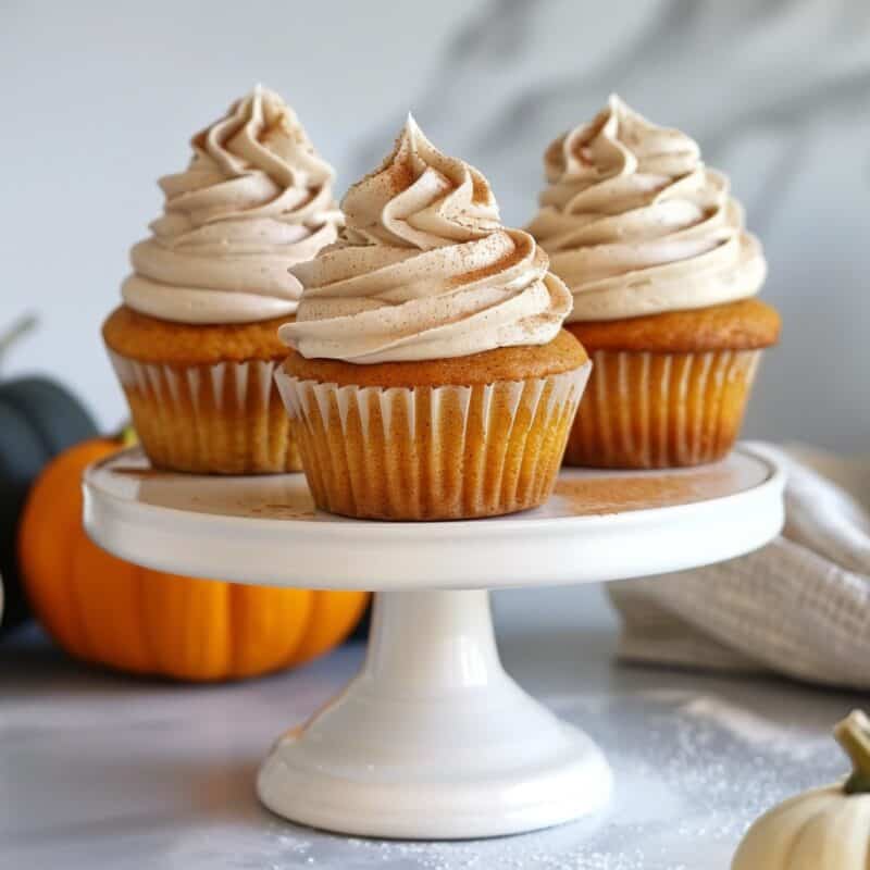 Three pumpkin cupcakes topped with generous swirls of cinnamon cream cheese frosting, displayed on a white cake stand, with small pumpkins in the background.