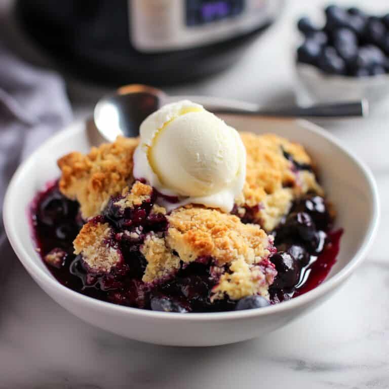 A close-up of Slow Cooker Blueberry Cobbler served in a white bowl, topped with vanilla ice cream, with fresh blueberries and a slow cooker in the background.