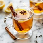 Two clear glass mugs of slow cooker mulled cider, each garnished with a slice of lemon, a cinnamon stick, and star anise, set on a marble surface with cloves and lemons in the background.