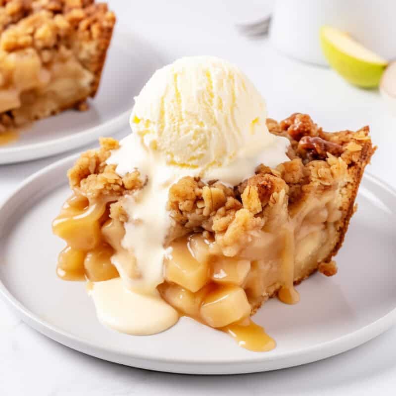 A close-up of a slice of apple dump cake on a white plate, topped with a scoop of vanilla ice cream. The cake features a golden-brown, crumbly topping with visible chunks of apple filling beneath. A drizzle of caramel sauce runs down the side, adding a glossy finish.