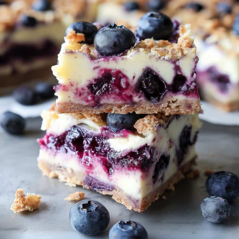 A square image featuring a stack of blueberry cream cheese bars with a thick, golden crust and blueberry-studded cheesecake filling, with fresh blueberries scattered on a rustic surface.