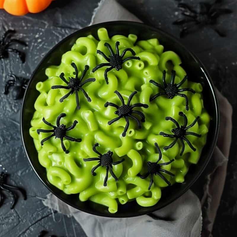 A black bowl containing green-colored mac and cheese, topped with black plastic spiders. The background is dark and decorated with Halloween-themed items, including small pumpkins and spiderwebs, enhancing the spooky presentation.