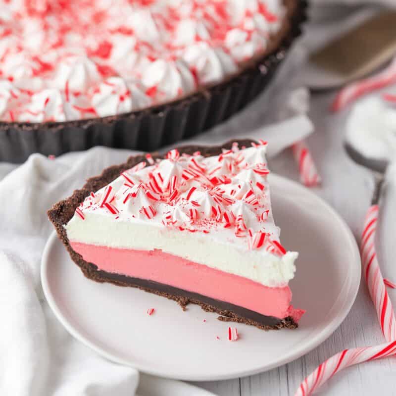 A slice of Layered Peppermint Pie on a white plate. The pie has a dark Oreo crust, a bright pink peppermint cream filling, and a layer of whipped cream topped with crushed peppermint candies. The entire pie is visible in the background.