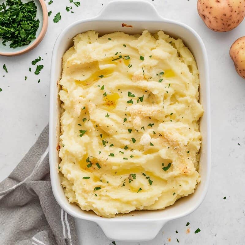 An overhead view of Potatoes Romanoff in a white baking dish. The dish is garnished with fresh parsley and has a golden crust, ready to be served as a rich and creamy side dish. Fresh potatoes and a small bowl of chopped parsley are placed beside the dish, indicating the fresh ingredients used.