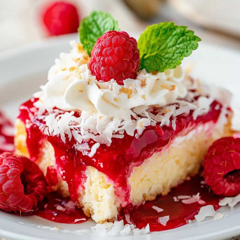 Square image of a Raspberry Zinger Poke Cake slice on a plate. The cake features a white cake base with a luscious layer of raspberry preserves and gelatin, topped with whipped cream and coconut flakes, and garnished with a raspberry and mint leaf.