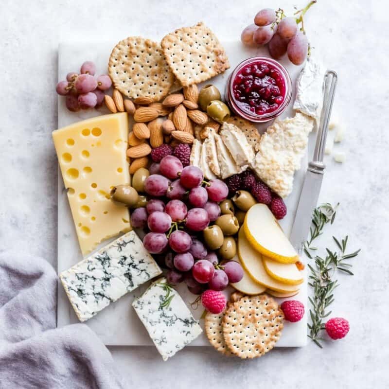 A beautifully arranged vegan Thanksgiving charcuterie board featuring an assortment of fresh grapes, raspberries, pear slices, various plant-based cheeses, nuts, crackers, bread slices, olives, and a small jar of cranberry sauce, all displayed on a white marble background.