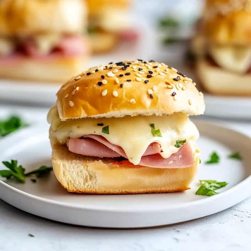 A single ham and Swiss cheese slider placed on a white plate, oozing melted cheese with fresh parsley garnish, highlighting the perfect blend of flavors for an easy appetizer or super bowl party food.