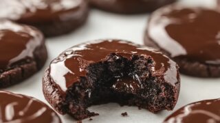 A batch of chocolate brownie cookies resting on parchment paper. One cookie has a large bite taken out, showing the gooey chocolate filling. The ganache on all cookies appears glossy and smooth, contrasting with the dark cookie base.