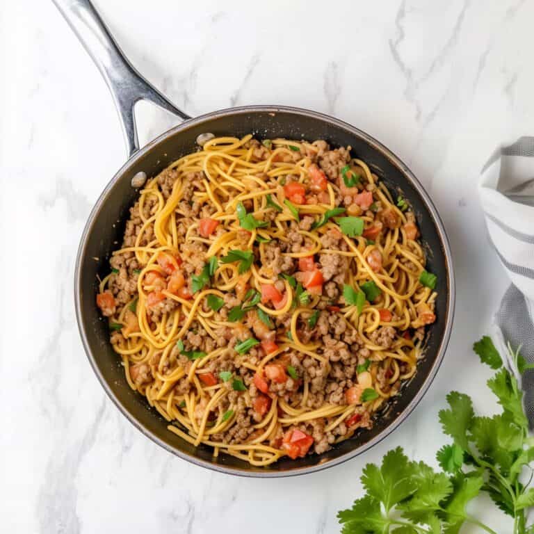 A skillet containing creamy taco spaghetti with ground beef, diced tomatoes, and fresh cilantro, set on a white marble surface with a gray napkin and a bunch of cilantro on the side.