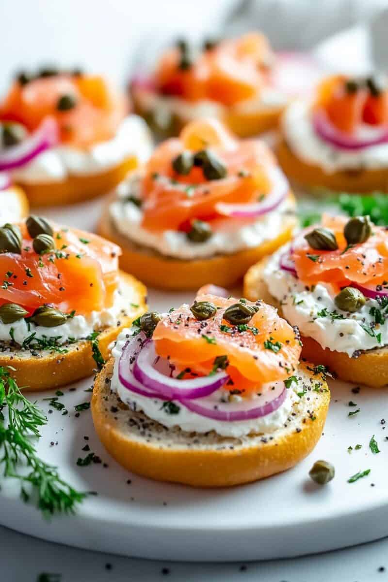 Elegant smoked salmon bagel bites arranged on a serving platter, topped with creamy spread, thinly sliced red onions, briny capers, and fresh dill, served on crispy bagel rounds.
