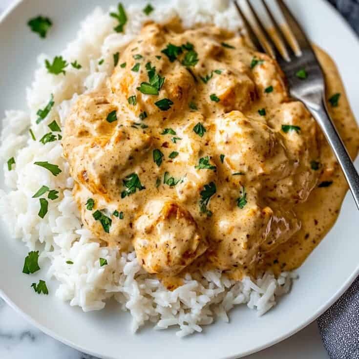 Overhead view of creamy smothered chicken served on a bed of white rice, topped with cilantro and photographed on a white plate with a fork.