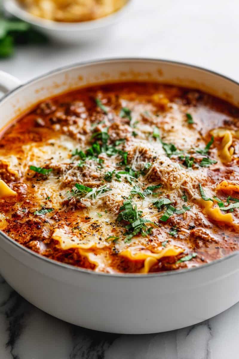 Close-up of Lasagna Soup in a white enamel pan topped with melted cheese, fresh basil, and lasagna noodles.