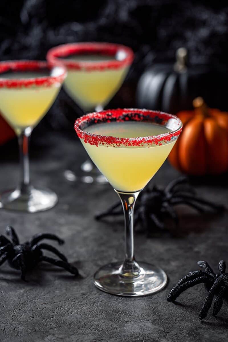 Three Haunted Candy Corn Cocktails in martini glasses, each with a striking red sugar rim, displayed among black spiders and pumpkins for Halloween ambiance.