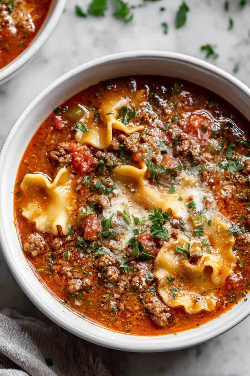 Top view of a bowl of Lasagna Soup with ground beef, tomatoes, lasagna noodles, and fresh herbs on a white background.
