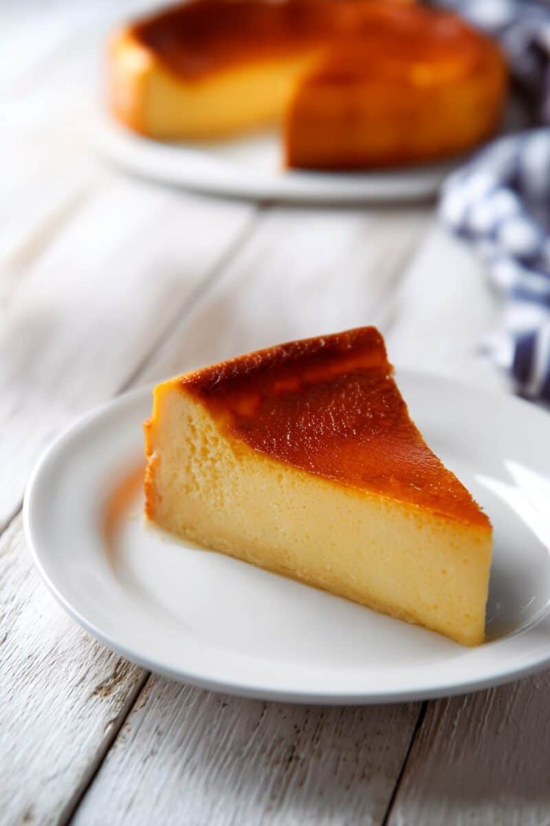 A slice of creamy pumpkin custard cake served on a white plate with the full cake in the background
