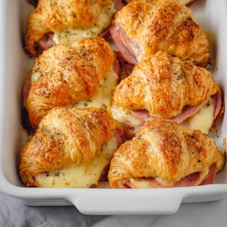 Golden baked ham and cheese croissants in a white ceramic baking dish, topped with herbs