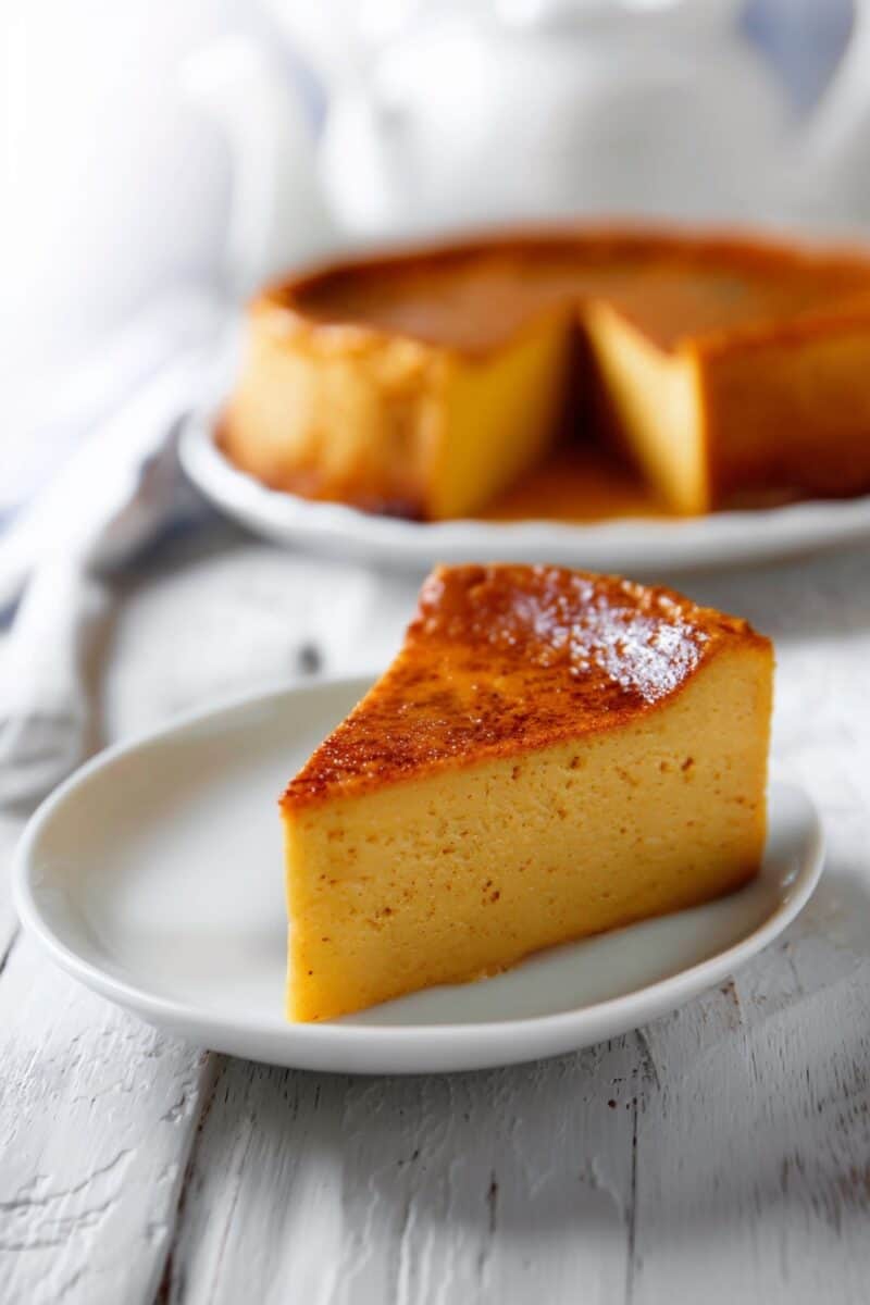 Silky slice of Pumpkin Custard Cake in front of the whole cake on a white wooden table
