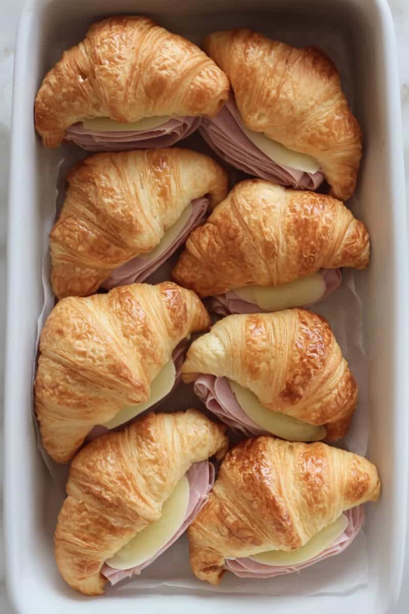 Unbaked ham and cheese croissants neatly arranged in a white casserole dish, ready to bake.