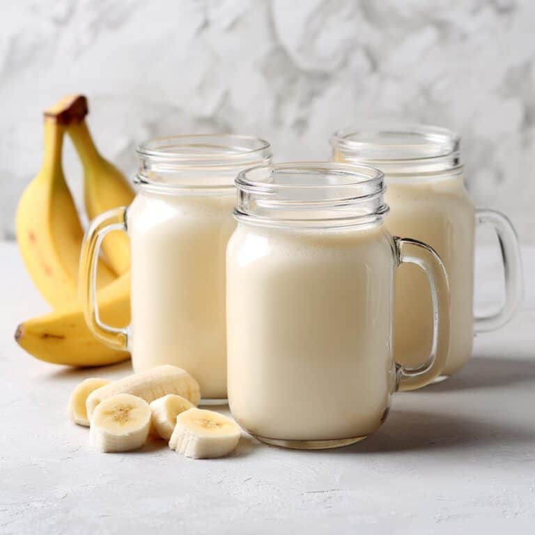 Banana milk served in clear mason jars with sliced bananas in front, styled on a white marble background.
