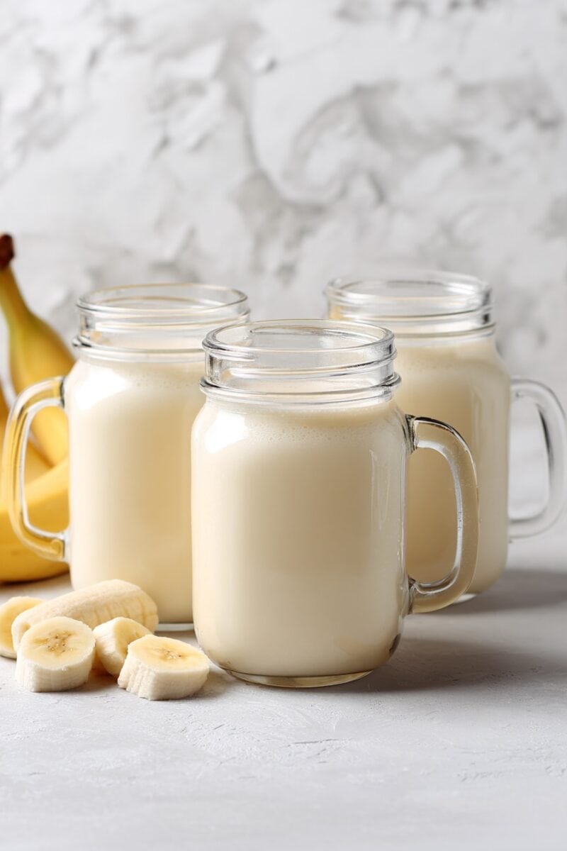 Three mason jars filled with banana milk on a marble surface, with sliced bananas and ripe bananas around them.