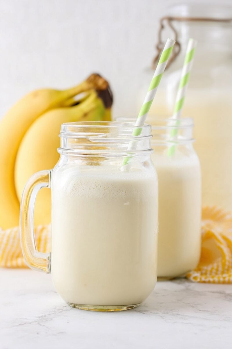 Two mason jars filled with creamy banana milk and green striped straws, with bananas in the background.